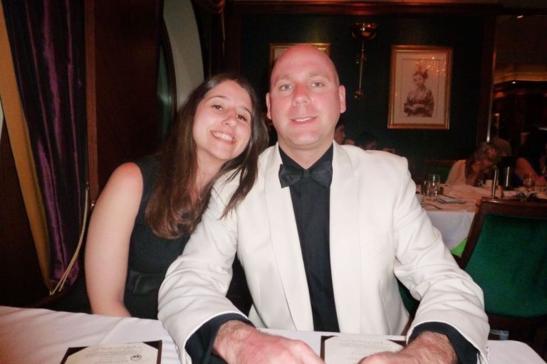 A smiling couple dressed in formal attire seated at a dining table during Royal Caribbean's formal night, with a menu in front of them and a classic portrait hanging on the wall in the background.