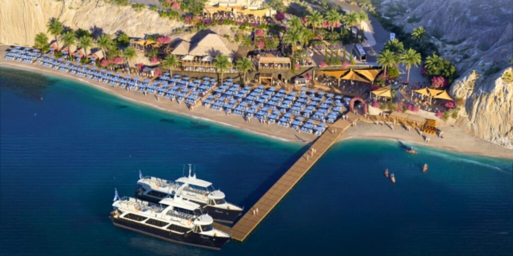 Aerial render of the upcoming Royal Beach Club Santorini, showing sun loungers shaded by umbrellas along a golden beach, surrounded by palm trees and bougainvillea. The design includes open-air dining areas, thatched-roof pavilions, and a wooden pier where yachts are docked against the island’s dramatic volcanic cliffs and clear Aegean water.