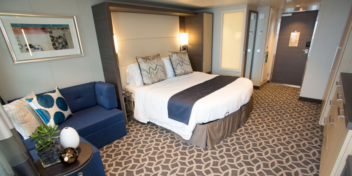 A spacious and elegantly appointed Royal Caribbean Junior Suite featuring a large bed with plush headboard and crisp white linens, a cozy blue sofa with decorative cushions, and a modern geometric-patterned carpet. The room is well-lit, inviting, and designed with a balance of comfort and style.