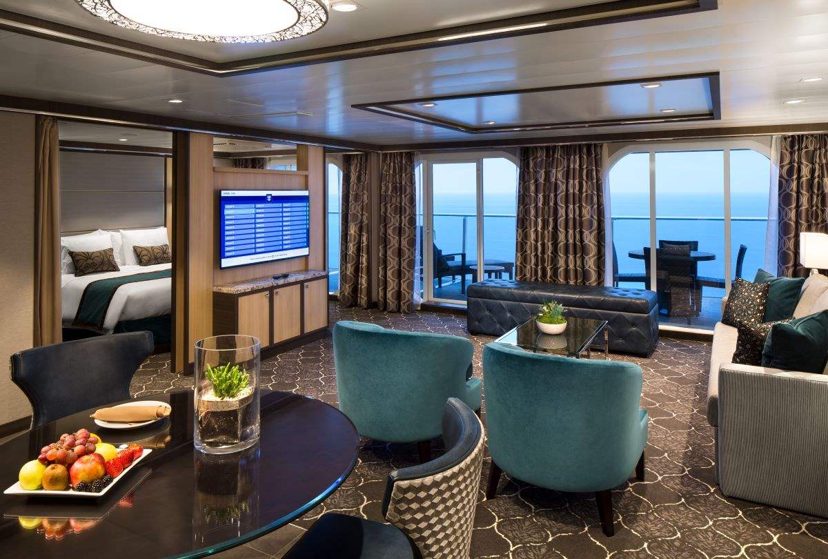 Opulent Owner's Suite on a Royal Caribbean cruise ship, showcasing a stylish living room with teal armchairs, a plush sofa, and a large flat-screen TV, adjacent to a bedroom area, all with panoramic ocean views through floor-to-ceiling windows.