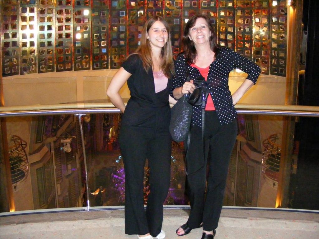 A cheerful mother and daughter posing together on Navigator of the Seas, standing by the railing with a colorful geometric-patterned wall in the background, capturing a moment of vacation joy.