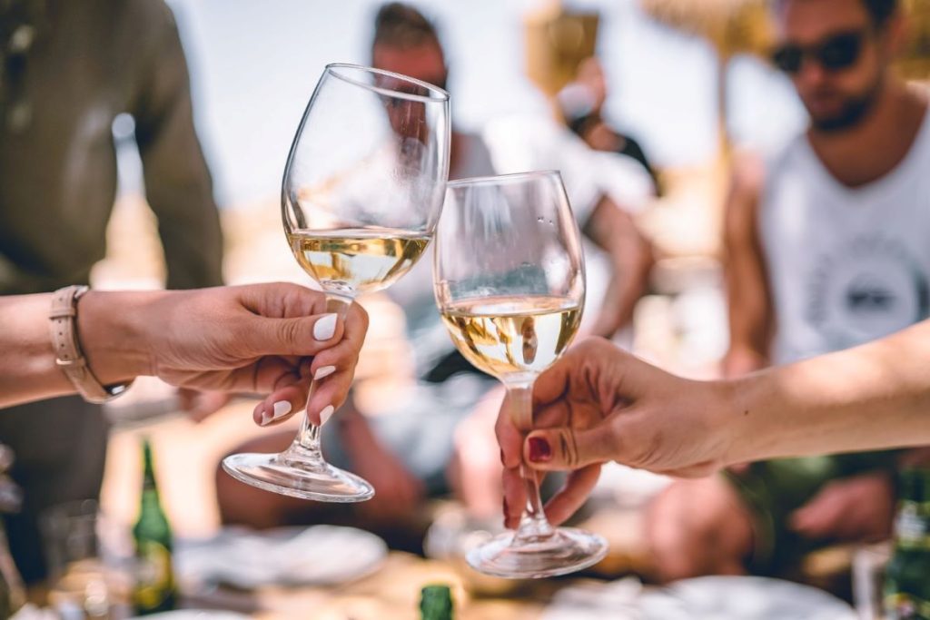 Close-up of two hands clinking glasses of white wine in a toast, with a blurred backdrop of a social gathering, possibly on a Royal Caribbean cruise, evoking a sense of celebration and leisure.
