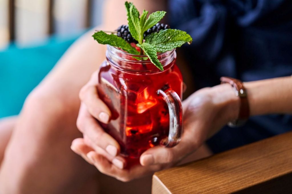 Close-up of a person's hands holding a mason jar filled with a vibrant red cocktail garnished with fresh mint and blackberries, offering a refreshing moment aboard a Royal Caribbean cruise.