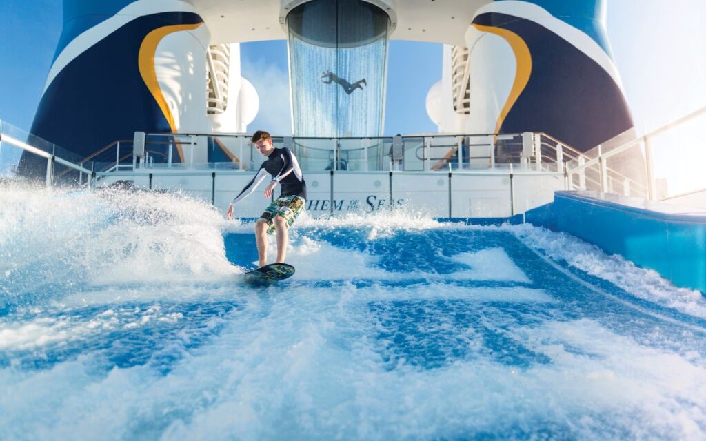 A person skillfully surfs on the Flowrider aboard Royal Caribbean's Anthem of the Seas, with the ship's distinctive smokestack in the background, under a bright blue sky