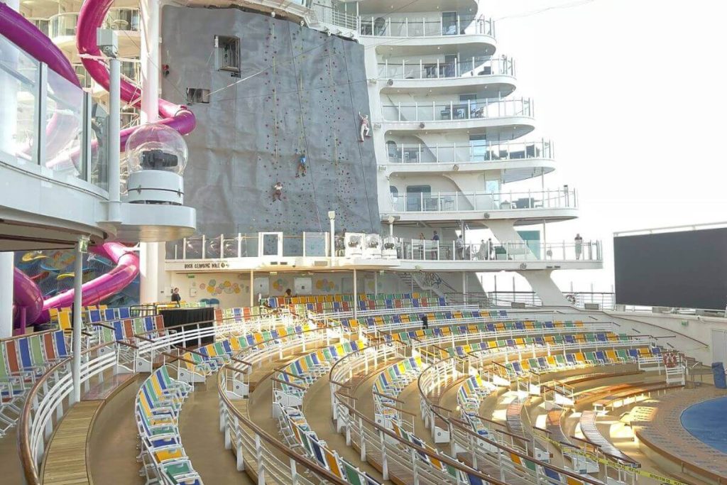 View of a bustling Royal Caribbean cruise ship deck featuring colorful waterslides, a rock climbing wall, and an outdoor amphitheater with rows of vibrant lounge chairs under a clear sky.