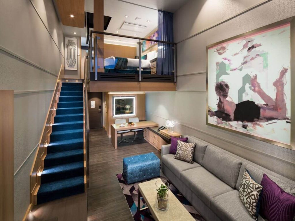 Elegant and modern Royal Caribbean suite interior, featuring a cozy living area with a gray sofa and vibrant cushions, an artistic wall painting, and a sleek staircase leading to an upper-level bedroom.