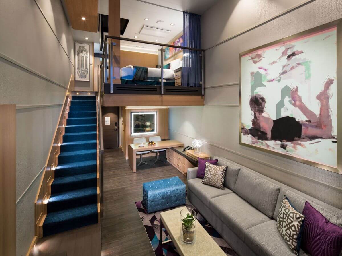 Elegant and modern Royal Caribbean suite interior, featuring a cozy living area with a gray sofa and vibrant cushions, an artistic wall painting, and a sleek staircase leading to an upper-level bedroom.