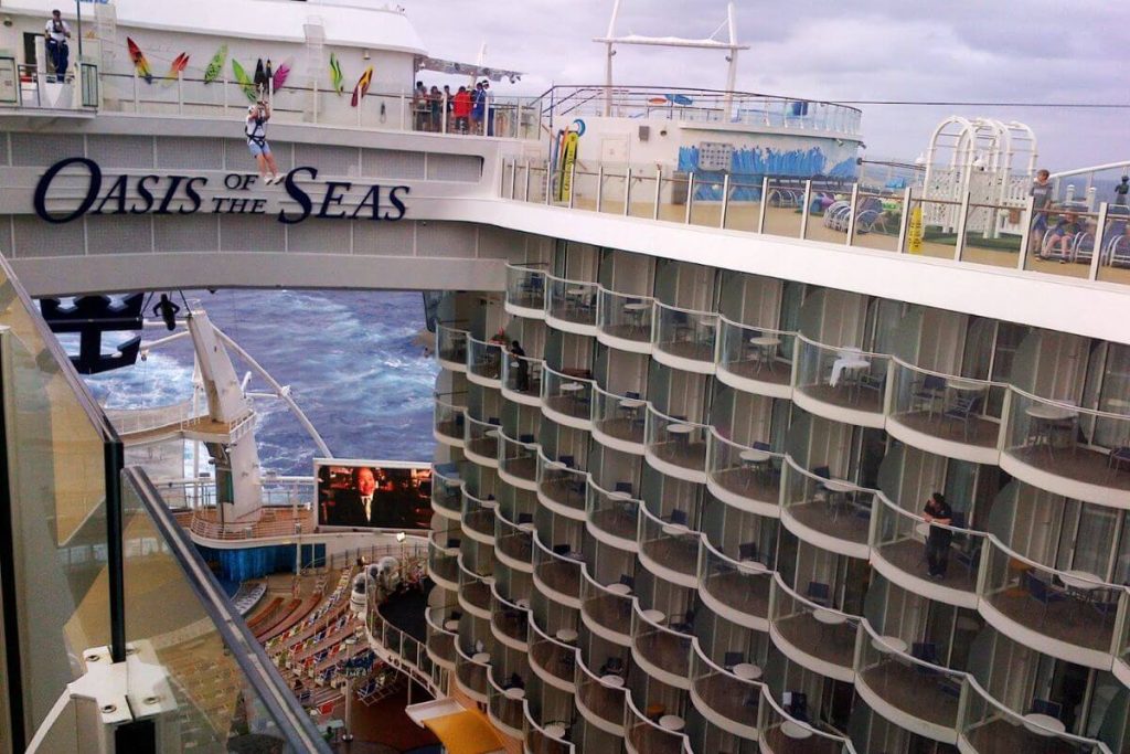 View of the Oasis of the Seas cruise ship featuring an adventurous guest ziplining above the boardwalk neighborhood, with the ship's multi-level balconies and an outdoor theater in view.