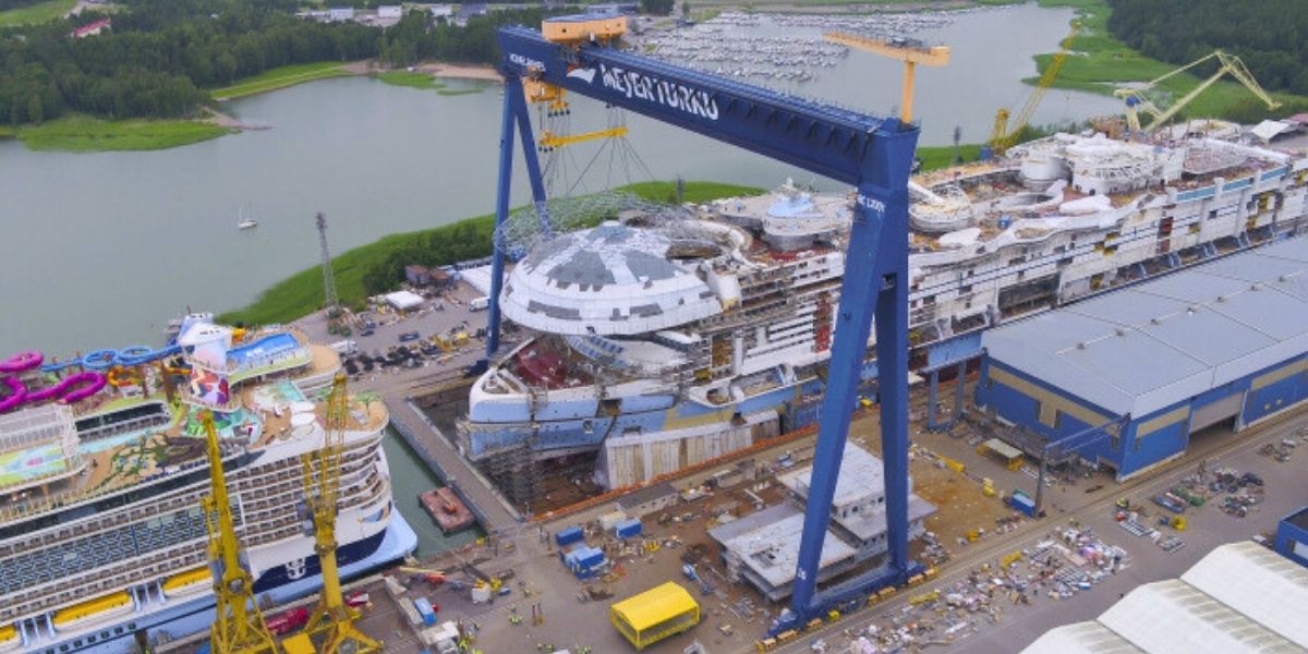 Aerial view of the Royal Caribbean cruise ship Utopia of the Seas under construction at the Meyer Turku shipyard in Finland. A massive blue crane labeled "MEYER TURKU" hoists a large circular structure onto the ship’s deck, surrounded by construction equipment, cranes, and a completed ship docked nearby.
