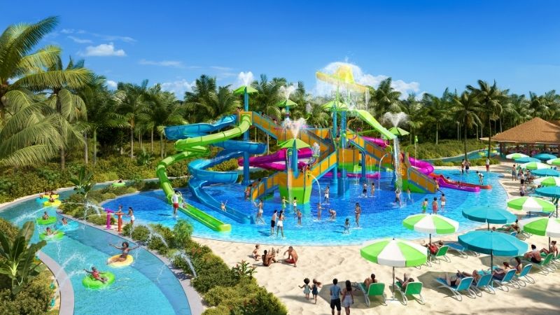 This image showcases Royal Caribbean’s upcoming Splashaway Bay at Perfect Day Mexico, set to debut in fall 2027. The colorful water play area features twisting slides, splash fountains, and a giant tipping bucket, all surrounded by palm trees, a lazy river, and shaded lounge chairs on sandy terrain.