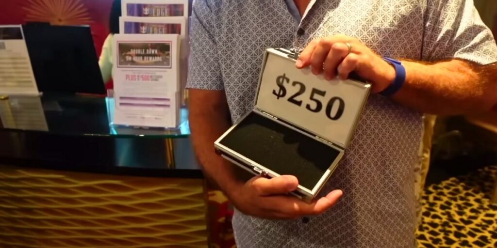 A man in a patterned shirt holds an open silver case displaying "$250" in bold black letters, likely representing casino or cruise promotional credit. The background includes a casino rewards desk with brochures, and the setting appears to be aboard a Royal Caribbean cruise.