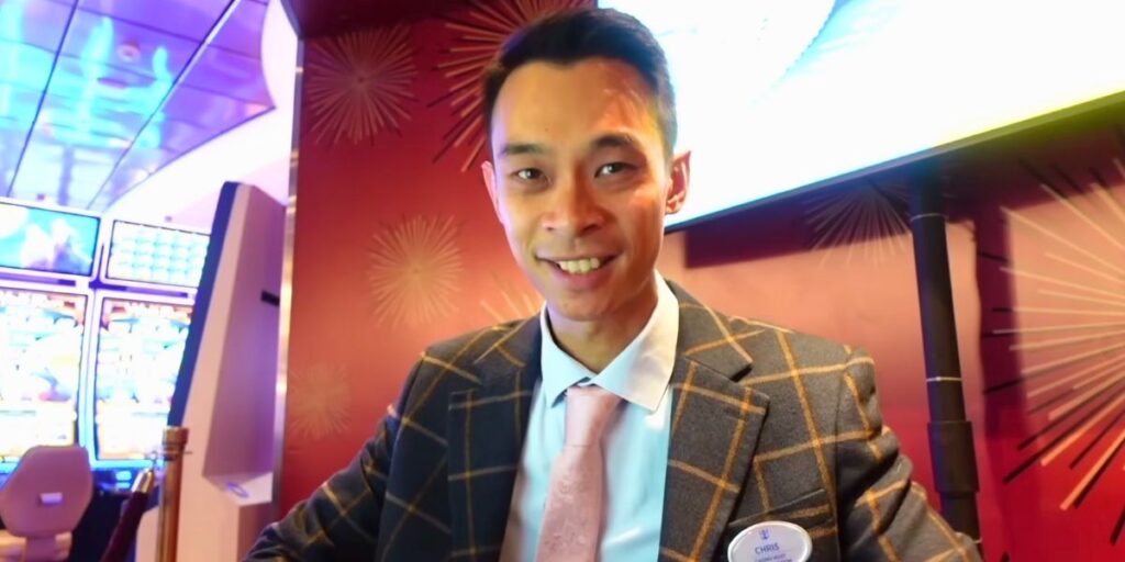 Chris Wong, a Royal Caribbean vlogger and casino host, smiles while standing in front of slot machines aboard a cruise ship. He wears a plaid blazer, pink tie, and name badge that reads “Chris,” set against a vibrant casino backdrop.