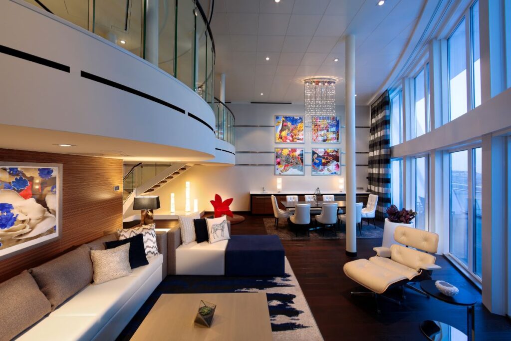 Interior view of the Royal Loft Suite on Anthem of the Seas featuring modern furnishings, a two-story glass window facade, and eclectic art pieces. The luxurious setting includes a plush seating area, contemporary lighting fixtures, and a sophisticated color palette of blues and neutrals.