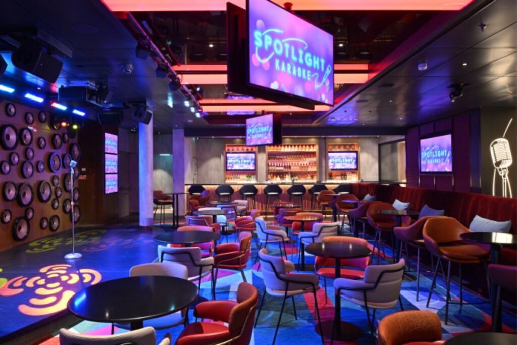 This image showcases the Spotlight Karaoke venue on Icon of the Seas, featuring a colorful and vibrant atmosphere. The space is furnished with modern chairs and tables, along with a performance stage set for karaoke enthusiasts. The neon lighting, large "Spotlight Karaoke" signs, and fun wall decor create an energetic and playful ambiance. The bar in the background adds to the lively experience, making it a perfect spot for guests to sing and enjoy the nightlife aboard the ship.