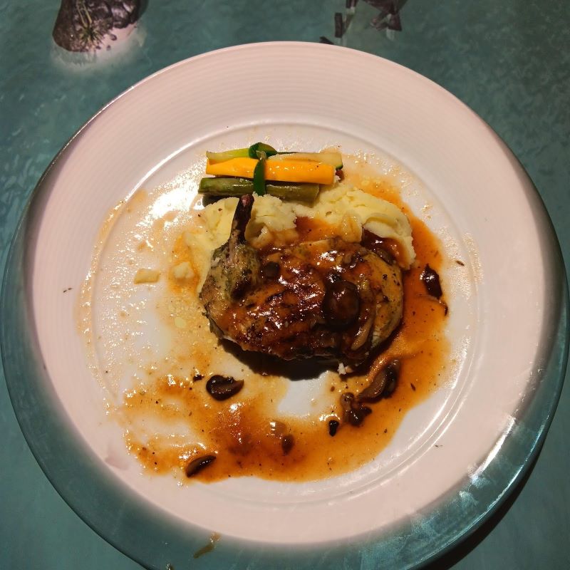A sautéed chicken breast is served on a white plate with a savory mushroom sauce, accompanied by creamy mashed potatoes and a garnish of sautéed vegetables. The plate is placed on a table with a subtle animated projection, adding a unique visual element to the dining experience at Le Petit Chef on the Celebrity Apex cruise ship.