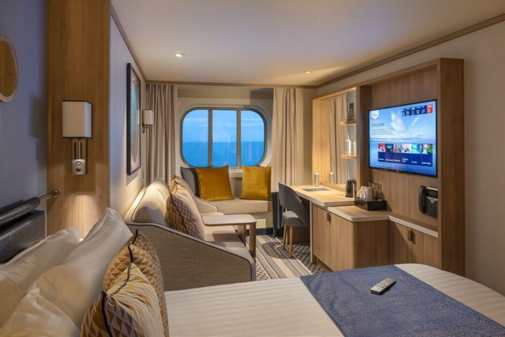 The image showcases a Sea View Cabin on the P&O Iona cruise ship. The cabin features a large round window with ocean views, a cozy sitting area with mustard-yellow cushions, and a comfortable bed with patterned pillows. The modern decor includes a flat-screen TV, wood-accented furnishings, and a neatly arranged desk area.