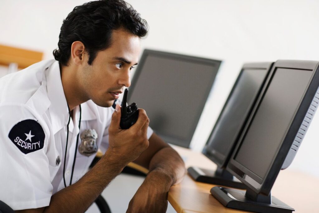 A security guard in a white shirt with a badge labeled 'SECURITY' attentively uses a walkie-talkie, with computer monitors in the background, illustrating a professional in communication.