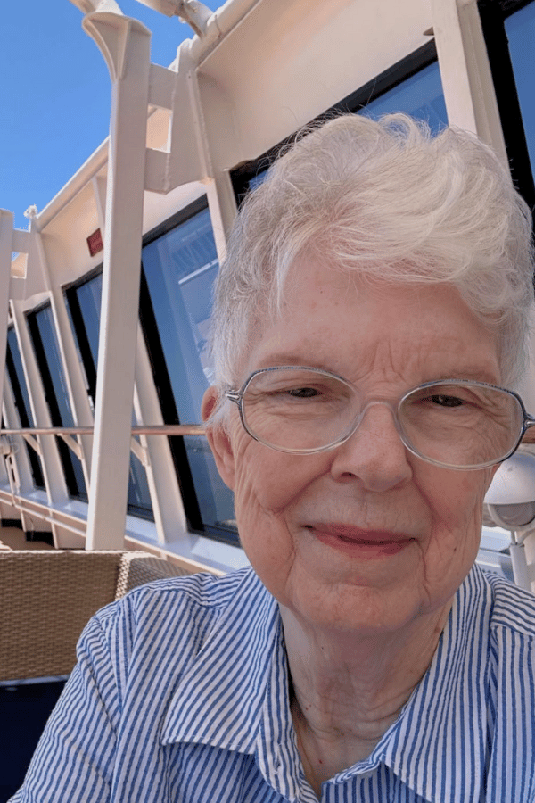 Sharon Lane, an older woman with short white hair and glasses, smiles while sitting on the deck of a cruise ship. She's wearing a light blue and white striped shirt, with the ship's railing and windows behind her under a clear blue sky.
