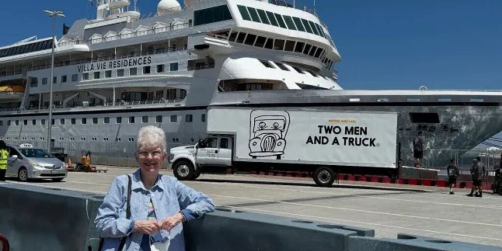 Elderly woman, identified as Sharon Lane, smiles while standing at port in front of the Villa Vie Residences cruise ship. A moving truck from "Two Men and a Truck" is parked nearby, suggesting preparations for long-term living aboard the ship.