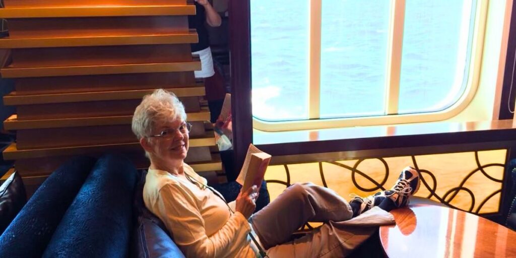 Sharon Lane, an elderly woman with short white hair and glasses, stands in front of a moving truck labeled "Two Men and a Truck" with the Villa Vie Residences cruise ship in the background. The image highlights her decision to sell all her belongings to live full-time on the cruise ship.