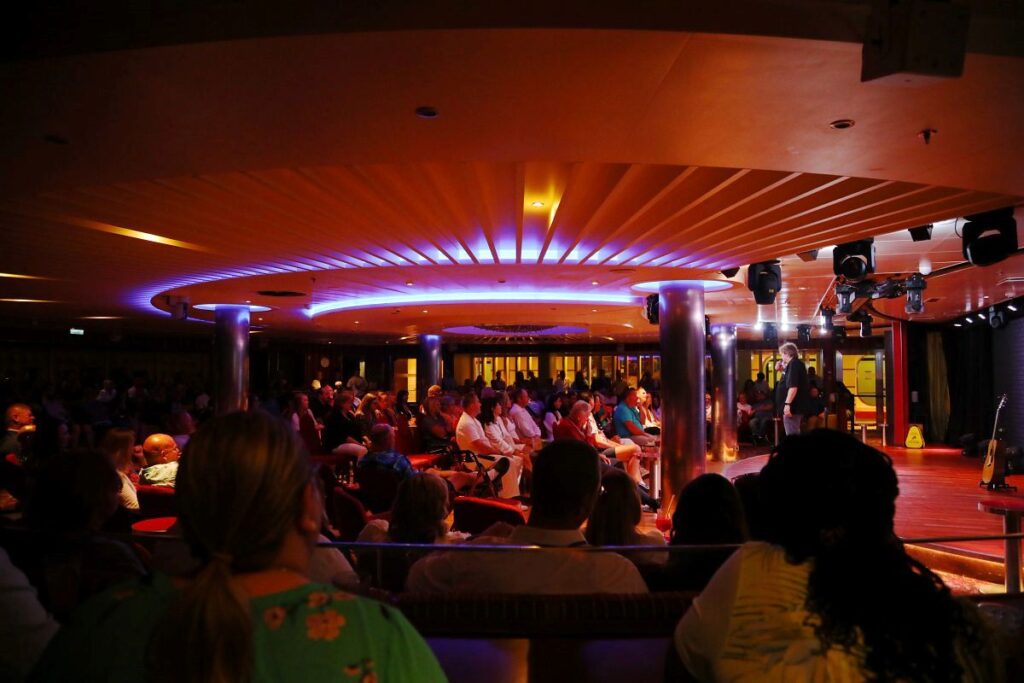 An evening show on a Carnival cruise ship features a performer on stage, entertaining a large audience seated in a cozy, dimly lit lounge. The room has a circular design with a vibrant ceiling illuminated in blue and purple hues, creating an intimate and engaging atmosphere. The audience appears captivated, enjoying the live performance as part of their cruise experience.