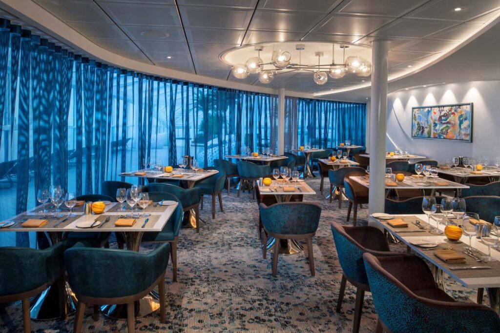 Silver Dining room on Spectrum of the Seas, featuring elegant teal and brown chairs, beautifully set tables with golden accents, and a serene ambiance enhanced by large windows draped with sheer blue curtains and contemporary art on the walls.