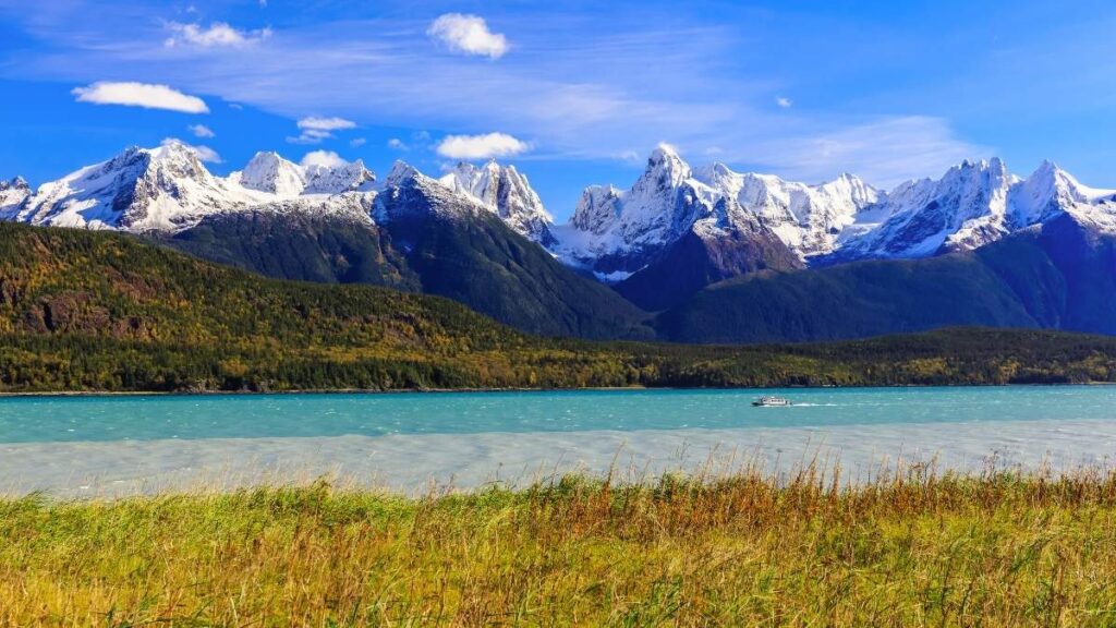 The image captures a scenic Alaskan adventure, a part of Carnival Cruise's shore excursions in Skagway, Alaska. It features a tranquil river with groups of adventurers paddling in inflatable rafts, surrounded by the majestic beauty of towering mountains and a stunning glacier in the distance. The lush greenery of the riverside foliage adds a pop of color to the serene landscape, illustrating a perfect blend of exhilarating activity and breathtaking nature.
