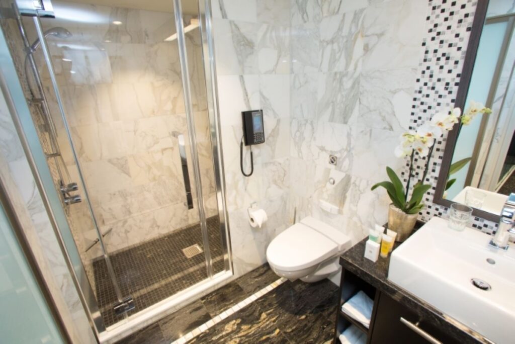 The luxurious restroom of the Sky Loft Suite on Anthem of the Seas showcases a spacious glass-enclosed shower, marble walls, and a sleek vanity area, complete with modern amenities and elegant orchid accents.
