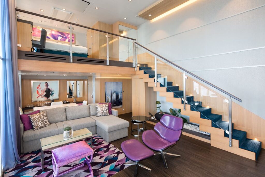 Interior view of the Sky Loft suite on a Royal Caribbean cruise ship, featuring a modern living area with a plush sectional sofa, vibrant carpet, and a sleek staircase leading to an upper-level bedroom, bathed in natural light from panoramic windows.