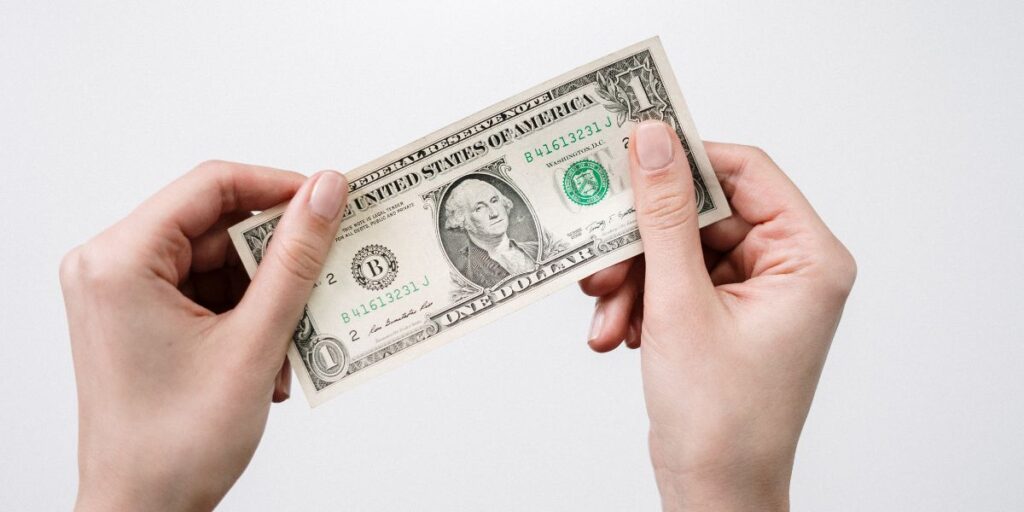Two hands holding a one-dollar bill against a white background, showcasing the front side of the currency with the portrait of George Washington.