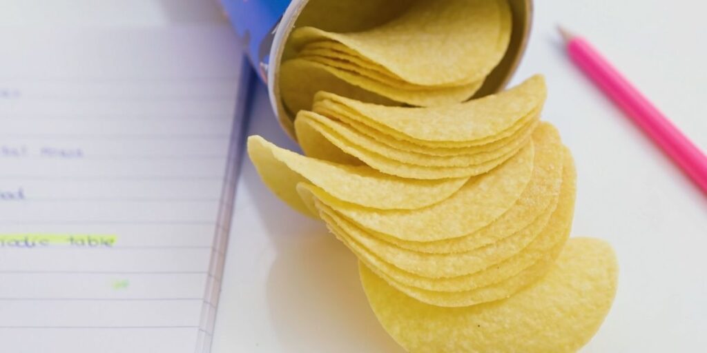 Close-up of a stack of uniform potato chips spilling out of a cylindrical can onto a white surface, with a pink pencil and a notebook containing highlighted notes visible nearby. The setup suggests a casual snacking moment during a study or work session.