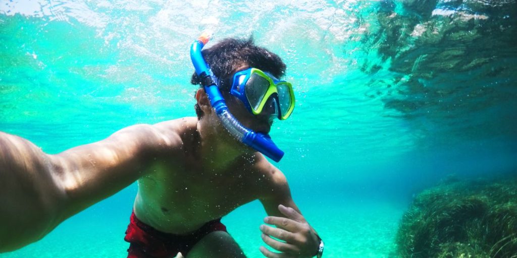 A person snorkeling in clear blue waters, wearing a blue snorkel and mask, with sunlight filtering through the surface creating a dappled effect on the water.