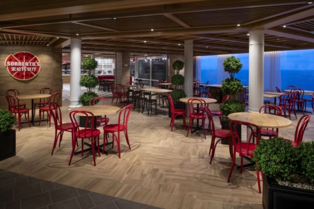 This image shows Sorrento's Pizzeria on Spectrum of the Seas, featuring a casual and inviting dining area with wooden tables and vibrant red chairs. The space is accented by greenery in the form of potted plants, creating a warm and welcoming atmosphere. The pizza-themed decor, with a large "Sorrento's" sign on the wall, adds a playful touch. Large windows in the background offer views of the ocean, enhancing the dining experience with scenic surroundings.