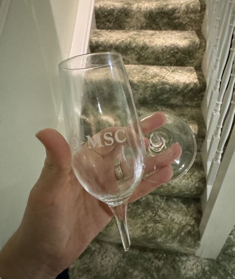 A hand holding a broken MSC-branded champagne flute, with the stem detached from the bowl, shown against a carpeted staircase in the background.
