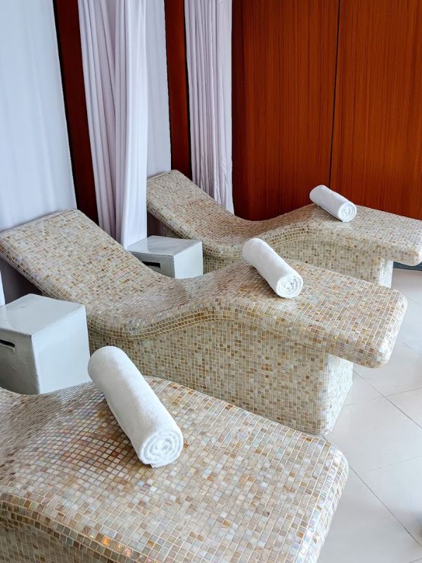 This image shows a serene spa area on the Oceania Mariner cruise ship. It features mosaic-tiled, curved loungers with rolled white towels placed on each seat, offering a relaxing environment. The loungers are spaced in front of wooden paneling, with white curtains providing a peaceful and calming atmosphere for spa-goers to unwind.