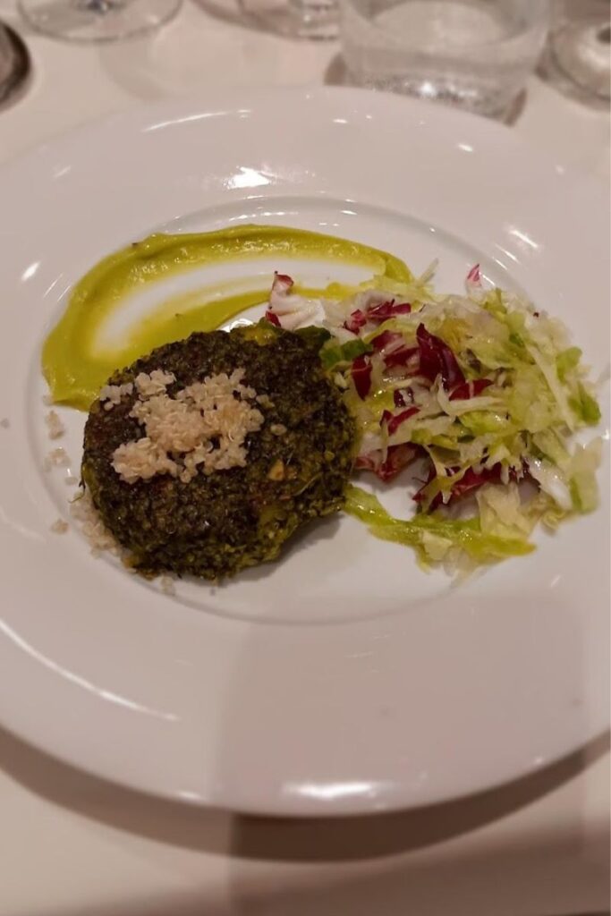 A gourmet vegetarian dish featuring a spinach, quinoa, and potato burger served alongside mesclun greens, garnished with an artistic swirl of avocado dressing on a white plate. The presentation is elegant, focusing on the textures and colors of the healthy ingredients.