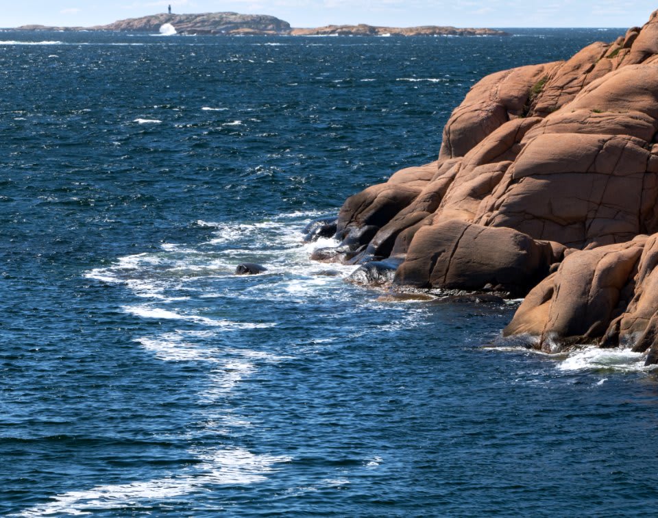 Waves hitting red granite sea cliffs along a rocky coastline, with a small island in the distance.