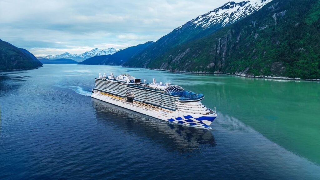 A majestic Princess Cruises ship sails through a breathtaking fjord, surrounded by towering, snow-capped mountains and lush green cliffs. The vessel's sleek white exterior, adorned with the signature blue wave design, contrasts beautifully with the deep blue and emerald waters below. The scene captures the serene and luxurious experience of an Alaskan or northern cruise adventure.