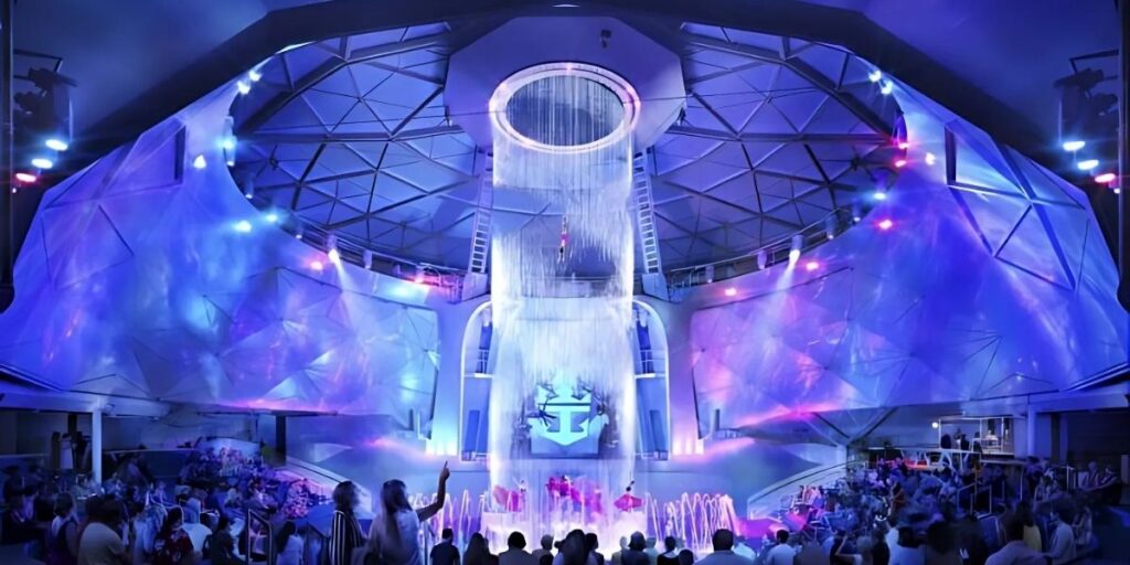 Royal Caribbean’s Star of the Seas Dome is dramatically lit in shades of purple and blue as an acrobat performs a mid-air stunt beneath a cascading waterfall, while a seated audience watches below. The futuristic venue features angular walls, spotlights, and a large Royal Caribbean crown-and-anchor logo illuminated in the background.
