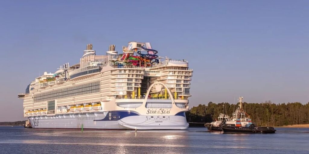 Royal Caribbean's massive "Star of the Seas" cruise ship is shown arriving with the help of a tugboat, surrounded by calm waters and a forested shoreline in the background. The ship's aft features bold blue and white detailing, yellow lifeboats, and a vibrant waterpark structure on the upper deck.