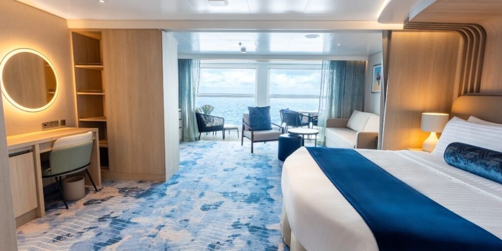 Spacious and modern cruise ship suite featuring a king-sized bed with a navy runner, a cozy sitting area by large ocean-facing windows, and a light wood vanity with a round illuminated mirror. The room’s decor blends soft blue carpeting with natural wood tones and sleek furnishings, creating a luxurious oceanfront retreat.