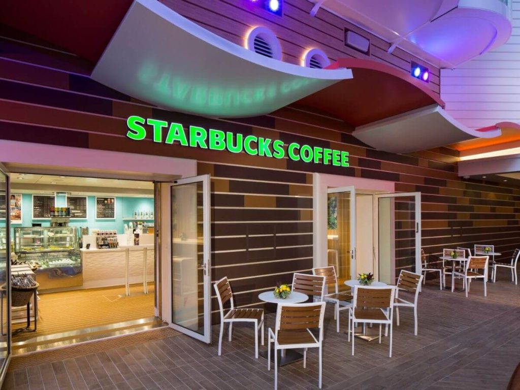 A Starbucks Coffee shop on Royal Caribbean's Harmony of the Seas, showcasing a bright and inviting outdoor seating area with white tables and wooden chairs, complemented by the signature green Starbucks sign above the entrance.