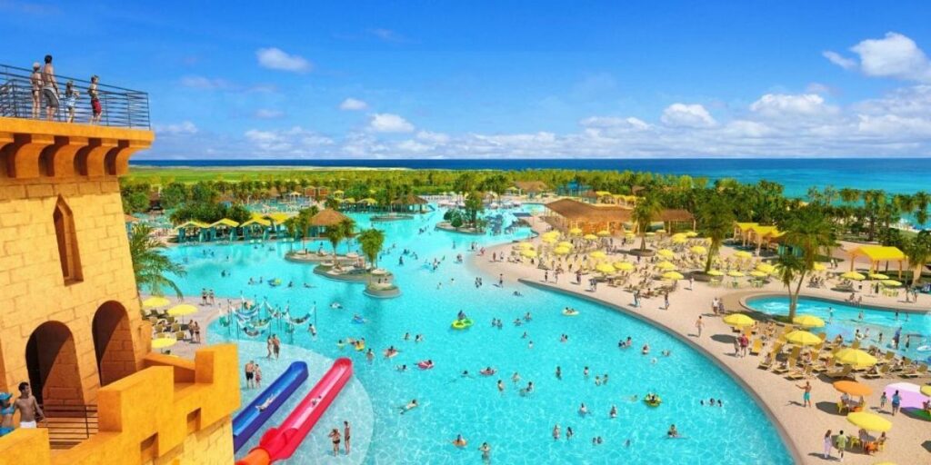 A wide aerial view of Starfish Lagoon at Celebration Key in The Bahamas, featuring a massive turquoise pool packed with guests, surrounded by yellow umbrellas, lounge chairs, and tropical palm trees. A yellow castle-like waterslide structure with red and blue slides stands at the edge, while the ocean stretches out in the background under a bright blue sky.
