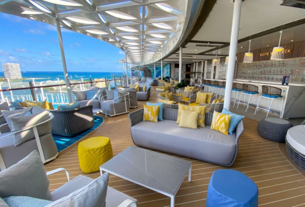 The Suite Sun Deck on Royal Caribbean's Wonder of the Seas offers a serene lounge area with modern gray and yellow accented furniture, overlooking the bright blue ocean under a geometric white canopy.