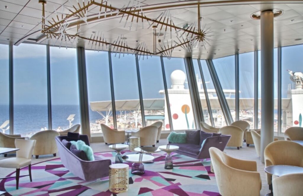 Chic suite lounge on Allure of the Seas with floor-to-ceiling windows offering a stunning sea view, eclectic purple-toned decor, contemporary art pieces, and a unique geometric ceiling installation creating a luxurious and inviting ambiance.