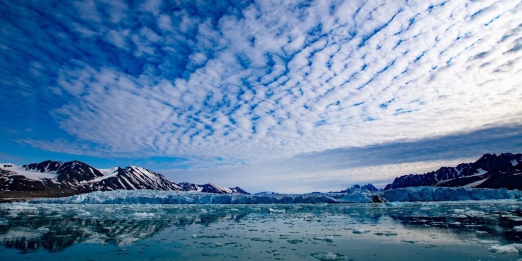 Panoramic view of snow-capped mountains and a glacier meeting icy Arctic waters in Svalbard, under a striking sky filled with rippling, textured clouds. Small chunks of ice float on the calm surface, mirroring the rugged landscape.