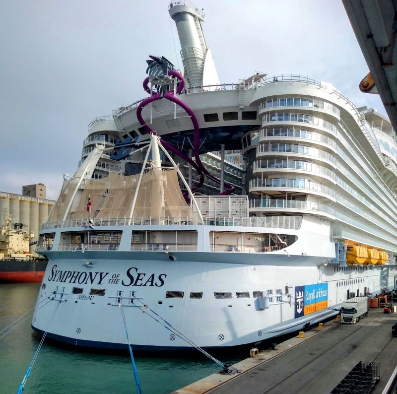 Docked at port, the Royal Caribbean Symphony of the Seas presents its stern, featuring a purple slide and multiple decks, against the industrial backdrop of a harbor, with mooring lines securing the vessel.