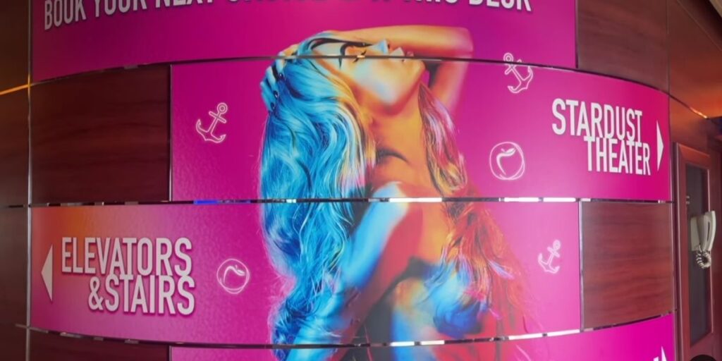 A bright pink wall sign aboard the Temptation Cruise featuring a stylized image of a woman with colorful hair and directions to the Stardust Theater and elevators.