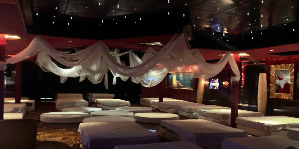 The Temptation Cruise playroom shown empty before opening, featuring low lighting, draped white fabric, and rows of circular and rectangular beds.