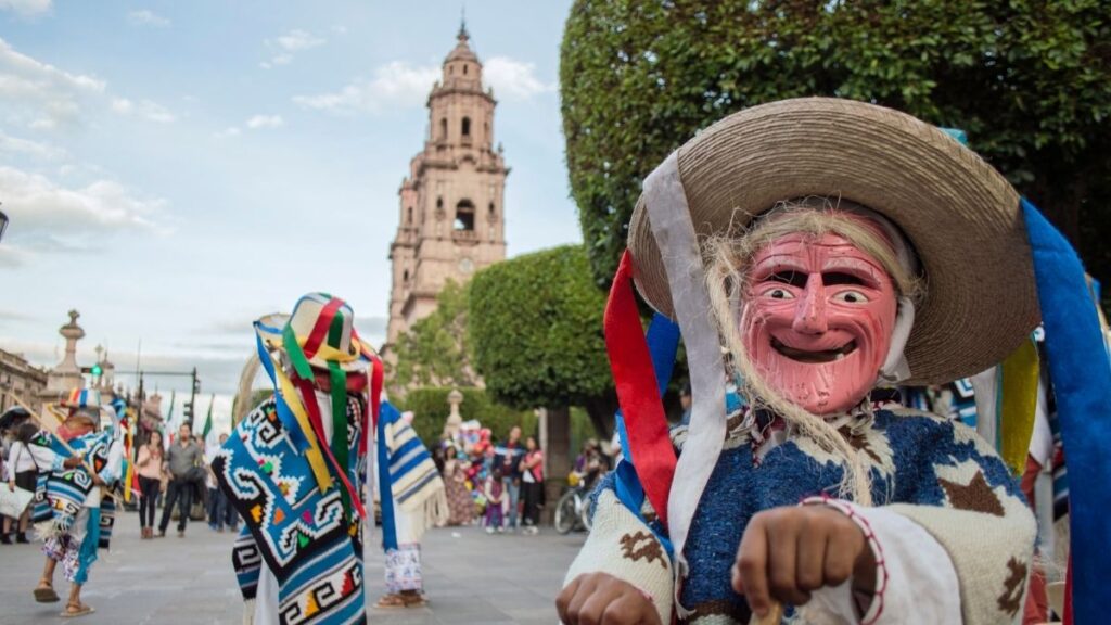 A vibrant cultural festival in a public square features a performer wearing a traditional wooden mask with exaggerated facial features, a woven sombrero adorned with colorful ribbons, and a blue and white poncho. Other dancers in similar attire, decorated with intricate patterns and bright ribbons, move energetically in the background. A historic church tower stands tall behind the festivities, adding to the rich cultural ambiance of the scene.